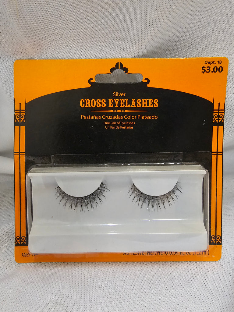 Packaged pair of silver cross eyelashes in a white plastic tray on an orange and black retail card, photographed against a white fabric background.