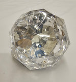A faceted clear glass or acrylic decorative object with a rounded, diamond-like shape, photographed on a light fabric background.