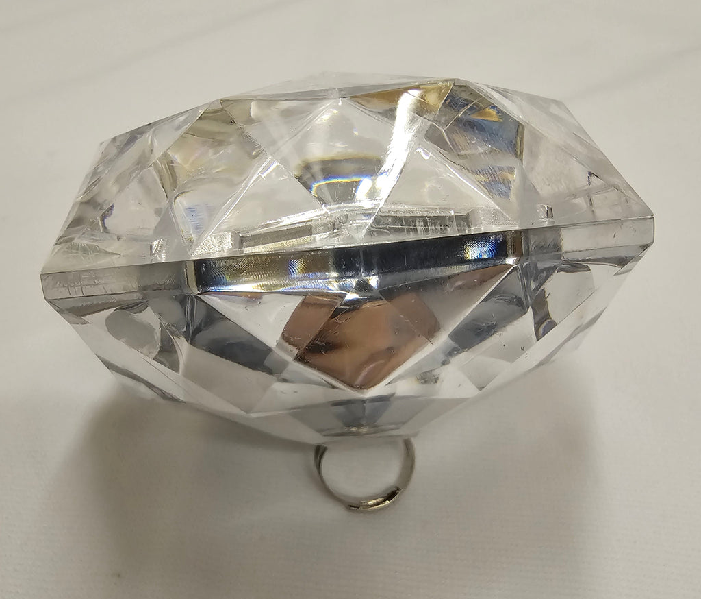 A clear faceted gemstone-shaped ring holder or decorative stand with a small metal ring at the base, photographed on a white fabric background.