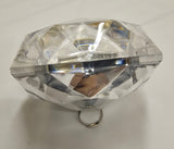 A clear faceted gemstone-shaped ring holder or decorative stand with a small metal ring at the base, photographed on a white fabric background.