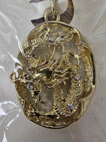A gold-toned oval pendant featuring an intricate dragon design with small clear rhinestone accents, shown hanging from a chain on a white textured surface.