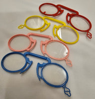 Six plastic novelty magnifying glasses in red, yellow, pink, and blue, arranged on a white textured fabric background.