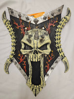 A decorative skull-shaped wall plaque or mask with a screaming face, mounted on a black shield-like backing with red flame motifs and yellow horn-like side elements, photographed against a light fabric background.