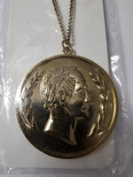 A round gold-toned pendant necklace featuring a profile portrait of a man surrounded by laurel branches, hanging from a chain on a white card inside clear plastic packaging.