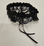 A black lace and velvet garter belt with a central fabric rose and two long ribbon ties, displayed on a light off-white textured background.