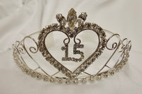 A silver wire tiara crown decorated with clear rhinestones, featuring a large heart outline with the number 15 in the center and a clear crystal flower at the top, displayed on a white fabric background.