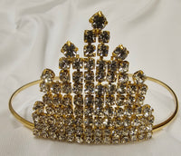 A gold-plated tiara-style headband adorned with multiple faceted clear rhinestones, set against a draped white fabric background.