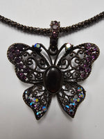 A butterfly-shaped pendant necklace with an antique bronze filigree design, featuring a central dark oval stone and purple rhinestone accents on the wings, suspended from a matching bronze chain against a plain light background.