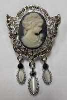 An ornate Victorian-style silver brooch featuring a central carved ivory cameo profile, surrounded by filigree detailing and three dangling teardrop charms with ivory and black elements, set against a plain light background.