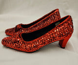 A pair of red sequined high-heeled shoes with pointed toes, displayed on a light gray fabric background.