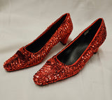 A pair of red sequined high-heeled pumps with pointed toes, displayed on a light gray fabric background.