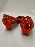 A pair of red sequined high-heeled shoes shown from the back on a white fabric background.