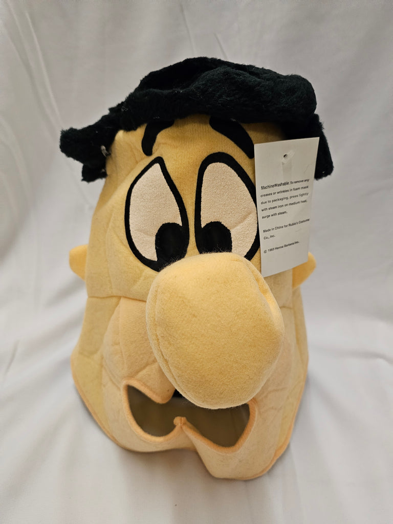 A plush novelty mask shaped like a cartoon character's face, featuring a yellow face, large cartoon eyes, a black hat, and a white product tag attached, set against a white fabric background.