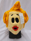 A plush costume mask resembling a cartoon or mascot face, with bright yellow hair, a cream face, black oval eyes, red lips, and a black display stand. A white product tag is attached to the side.