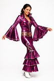 A woman modeling a shiny metallic magenta-purple 1950s-style flapper costume with long bell sleeves, a ruffled hem, and white high-heeled boots against a plain white studio background.