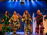 Four women perform on stage wearing shiny metallic ruffled outfits in green, gold, and purple tones against a blue curtain backdrop with a disco ball overhead.