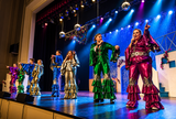 A group of performers in shiny, metallic ruffled outfits dancing on a brightly lit stage under blue and purple theatrical lights. The foreground performer wears a bright magenta-pink and silver outfit, while others wear green, blue, and silver costumes.