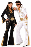 A man and a woman modeling Elvis-style costume jumpsuits against a plain white background. The woman wears a black flared jumpsuit with gold sequins and gold trim, and the man wears a white flared jumpsuit with gold sequins and gold trim.