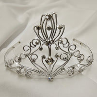 An ornate silver tiara crown with intricate filigree wirework and clear rhinestones, displayed on a white fabric background.