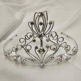 An ornate silver tiara crown with intricate filigree wirework and clear rhinestones, displayed on a white fabric background.